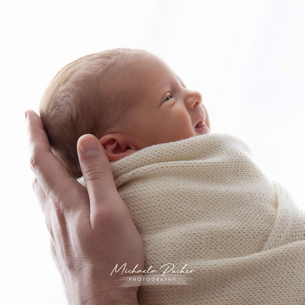 Vater hält ein eingewickeltes Neugeborenes sanft am Hinterkopf und das Baby lächelt mit geöffneten Augen.